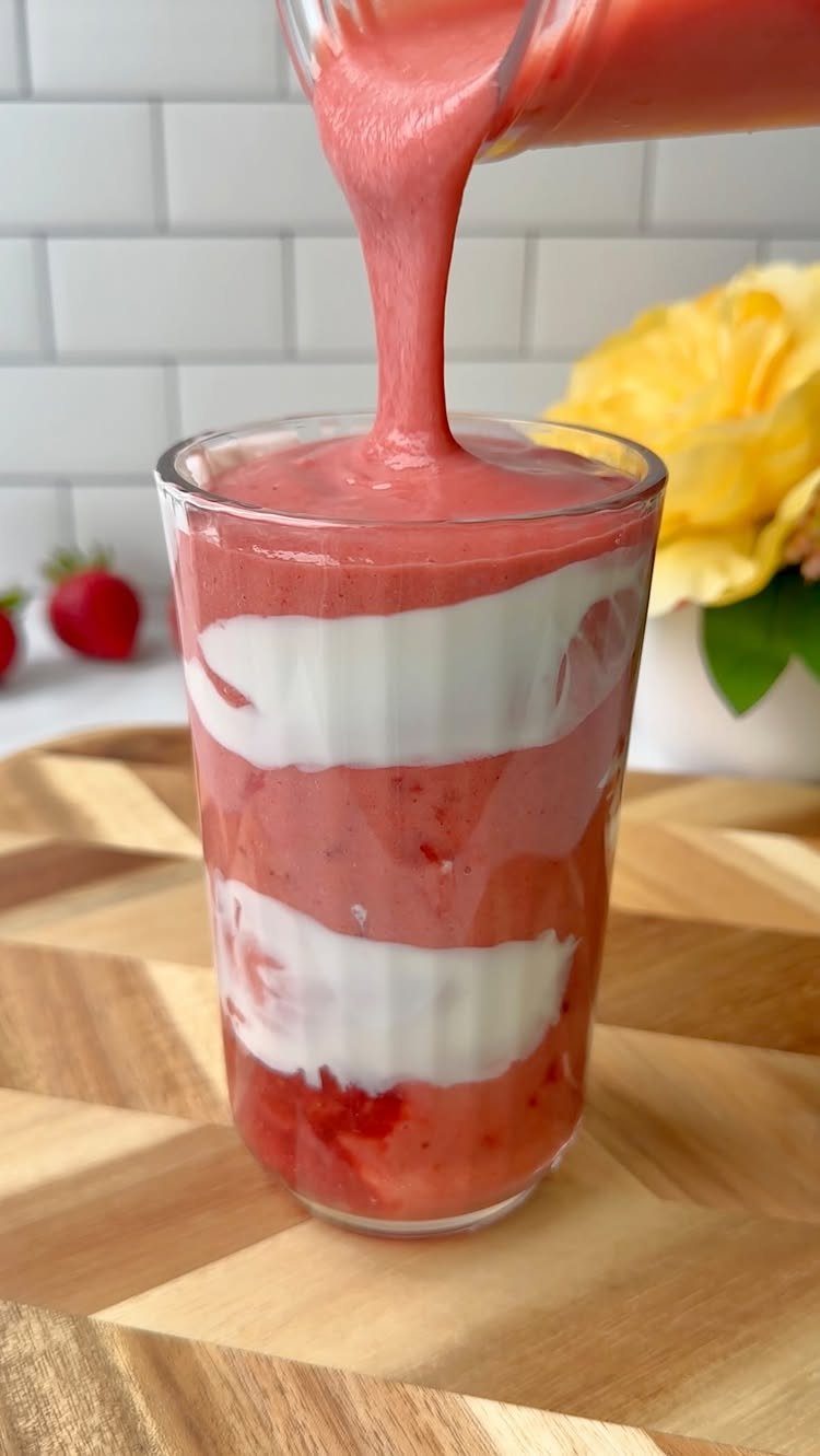 Pink strawberry smoothie being poured into a glass with white yogurt layers.