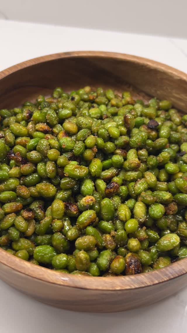 Roasted green edamame beans in a round wooden bowl on a white surface.