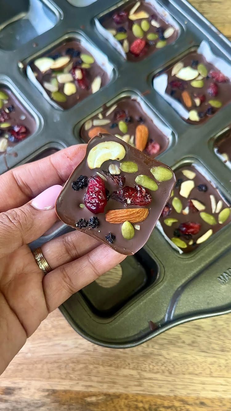 21 Healthy Mini Snacks for Lunchboxes, Parties and Busy Days 20 Chocolate bark piece with almonds, cashews, and dried berries held over a baking tray.