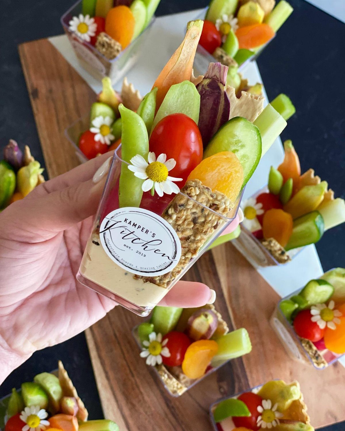 21 Healthy Mini Snacks for Lunchboxes, Parties and Busy Days 7 Hummus cup with sliced cucumbers, cherry tomatoes, and yellow peppers on a wooden board.