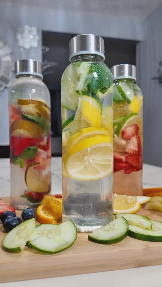 Fruit-infused water in glass bottles with fresh lemon, cucumber, and strawberries on wood.