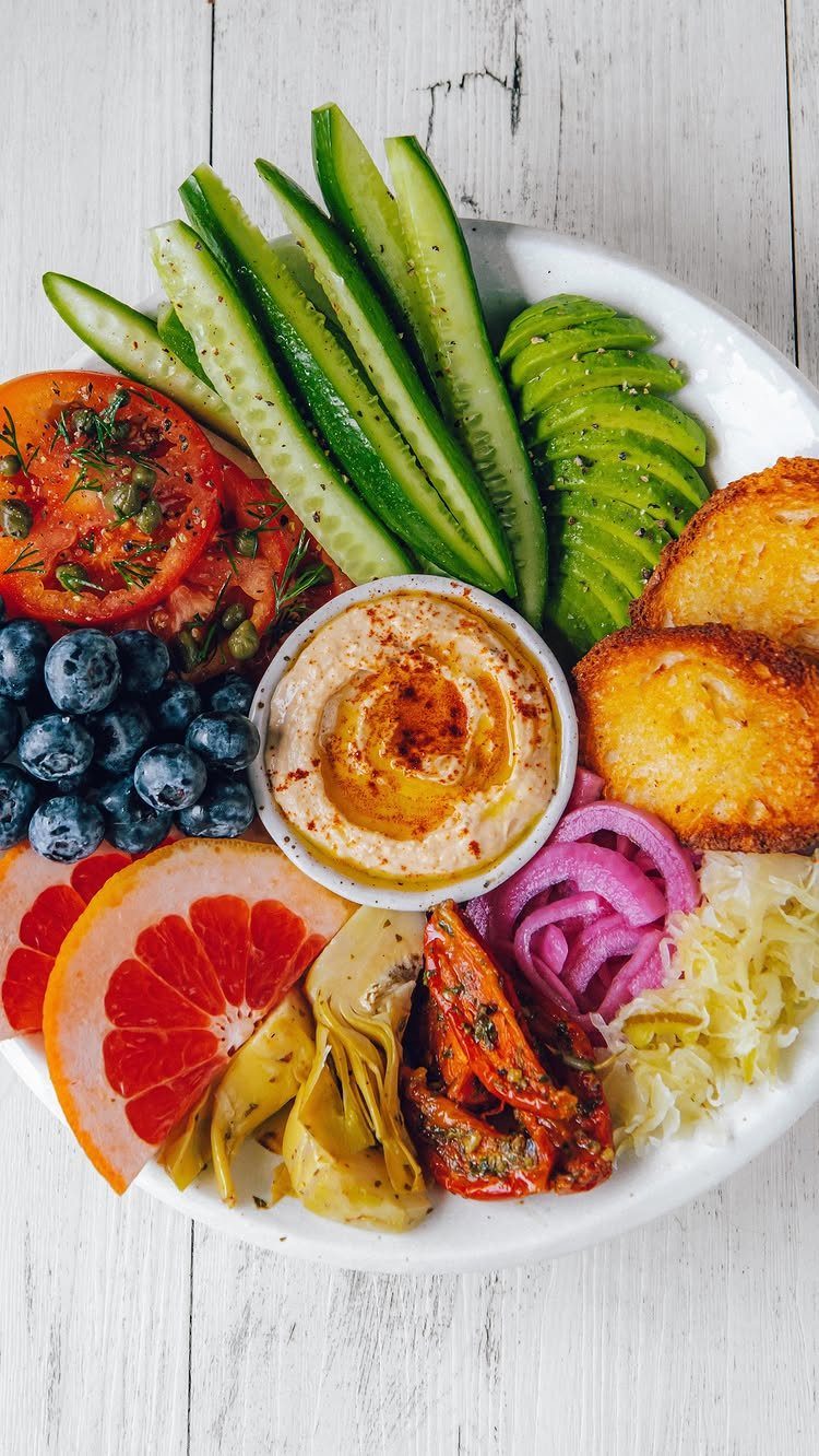 Hummus bowl surrounded by cucumber, avocado, tomatoes, blueberries, and grapefruit on a platter