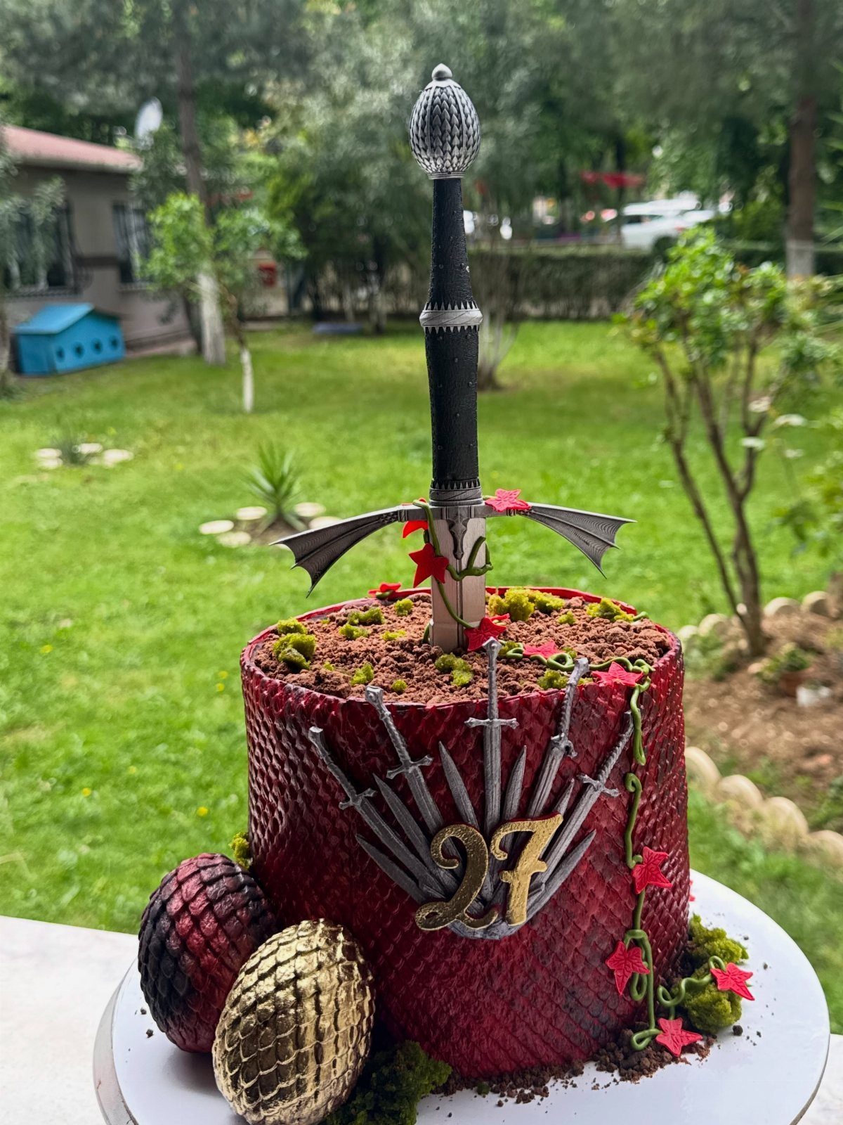 Red scale-textured cake with a large black sword, dragon eggs, and gold number 27.