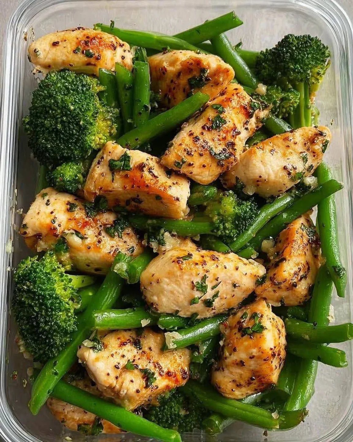 Chicken breast pieces with broccoli and green beans in a clear plastic container.