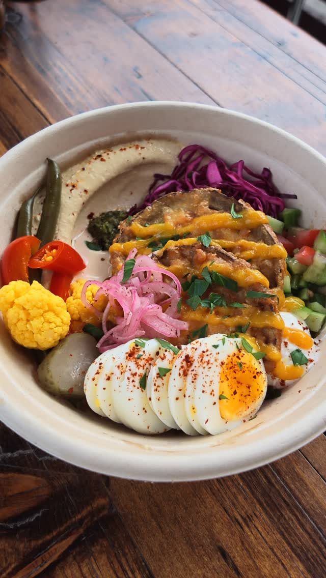 Hummus bowl with soft-boiled eggs, roasted eggplant, pickled onions, and colorful vegetables.