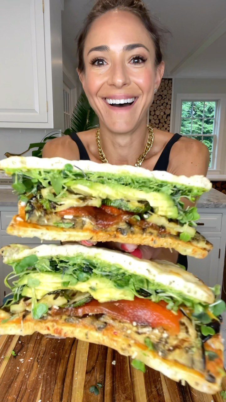 Veggie sandwich with avocado and roasted peppers held by a smiling woman.
