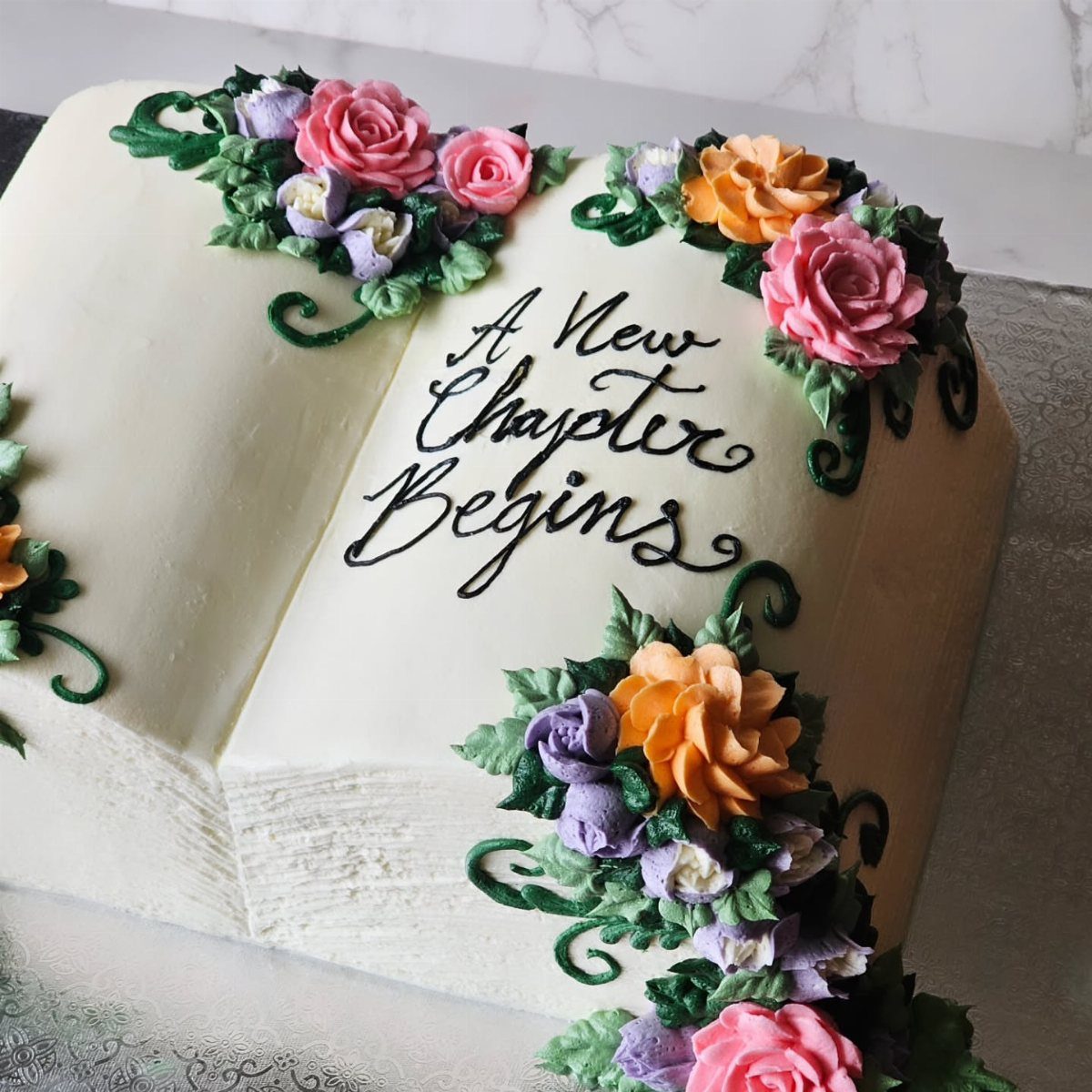 13 Open Book Cakes, from Fairytale Pages to Library Shelves 11 Open book cake with colorful frosting roses and text reading A New Chapter Begins.