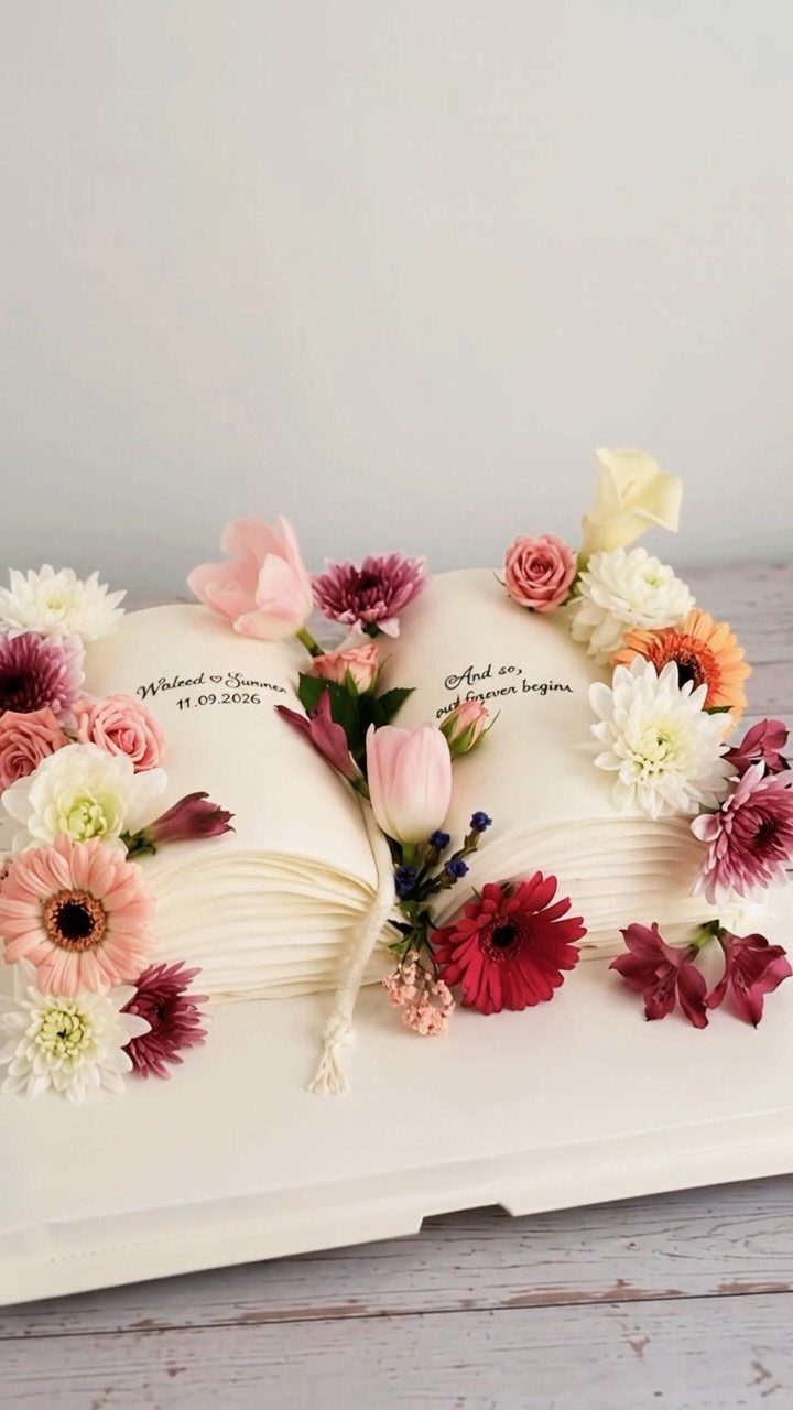 13 Open Book Cakes, from Fairytale Pages to Library Shelves 6 Open book cake with white frosting, decorated with fresh colorful flowers and cursive writing.