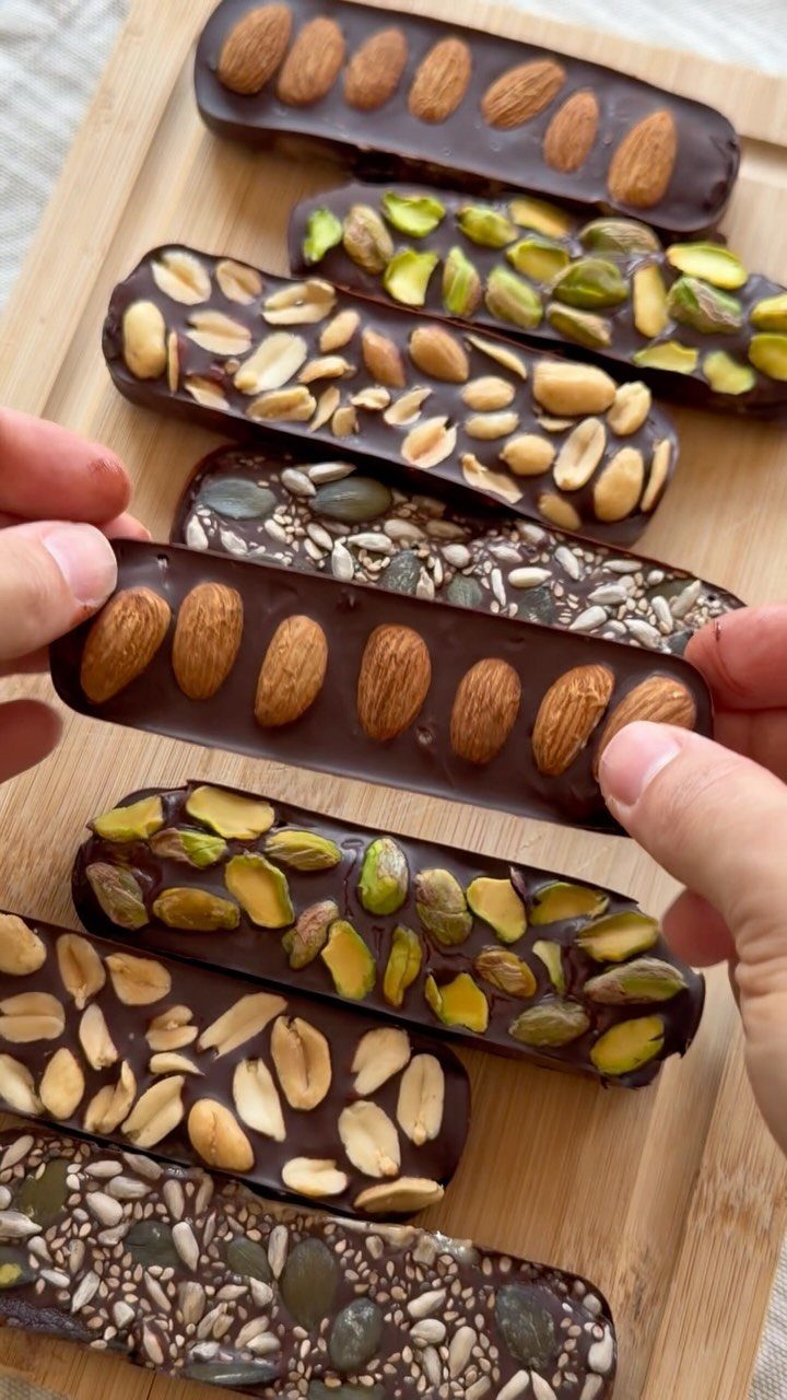 Dark chocolate bars topped with almonds, pistachios, and peanuts on a wooden board.