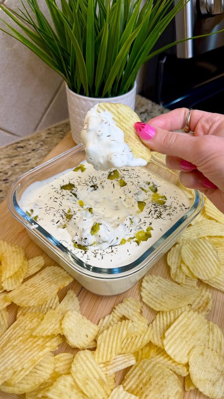Potato chip being dipped into a creamy white dill pickle dip in glass bowl.