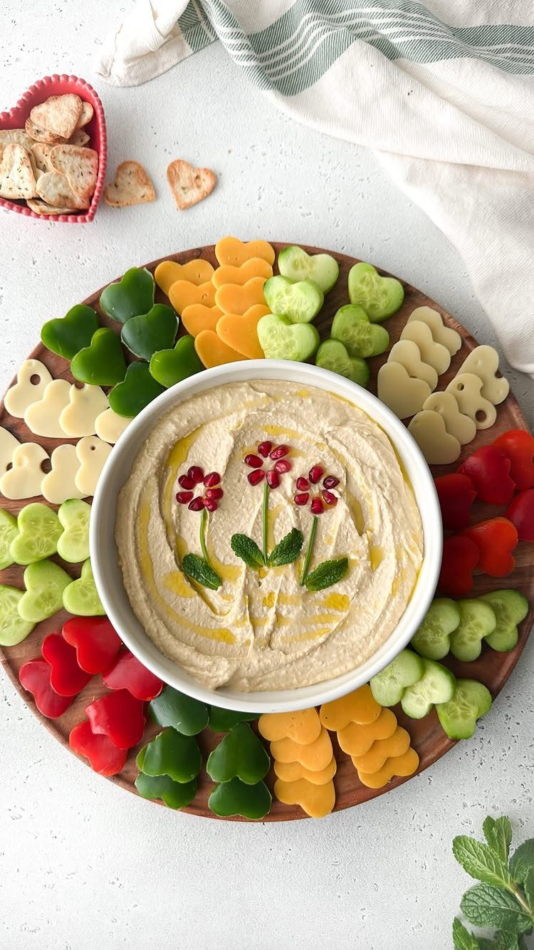 Hummus bowl surrounded by heart-shaped cheese, cucumber, and red pepper on wooden platter.