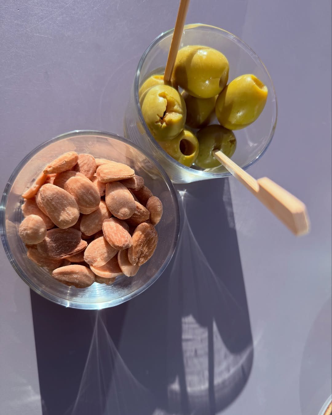 Green olives and brown almonds in two small glass bowls with wooden toothpicks.