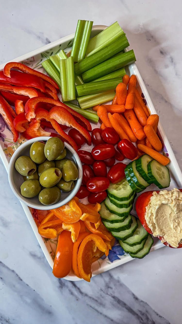 Vegetable platter with peppers, celery, carrots, cucumbers, olives, and hummus on marble.