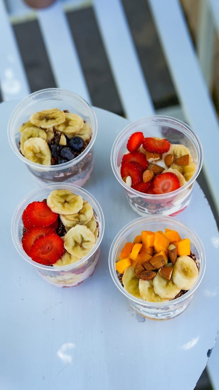 Four yogurt parfaits with granola, bananas, strawberries, and blueberries in plastic cups.