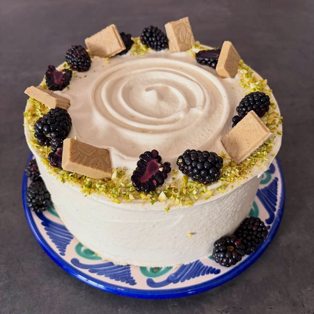 White frosted cake topped with blackberries, white chocolate squares, and crushed pistachios.
