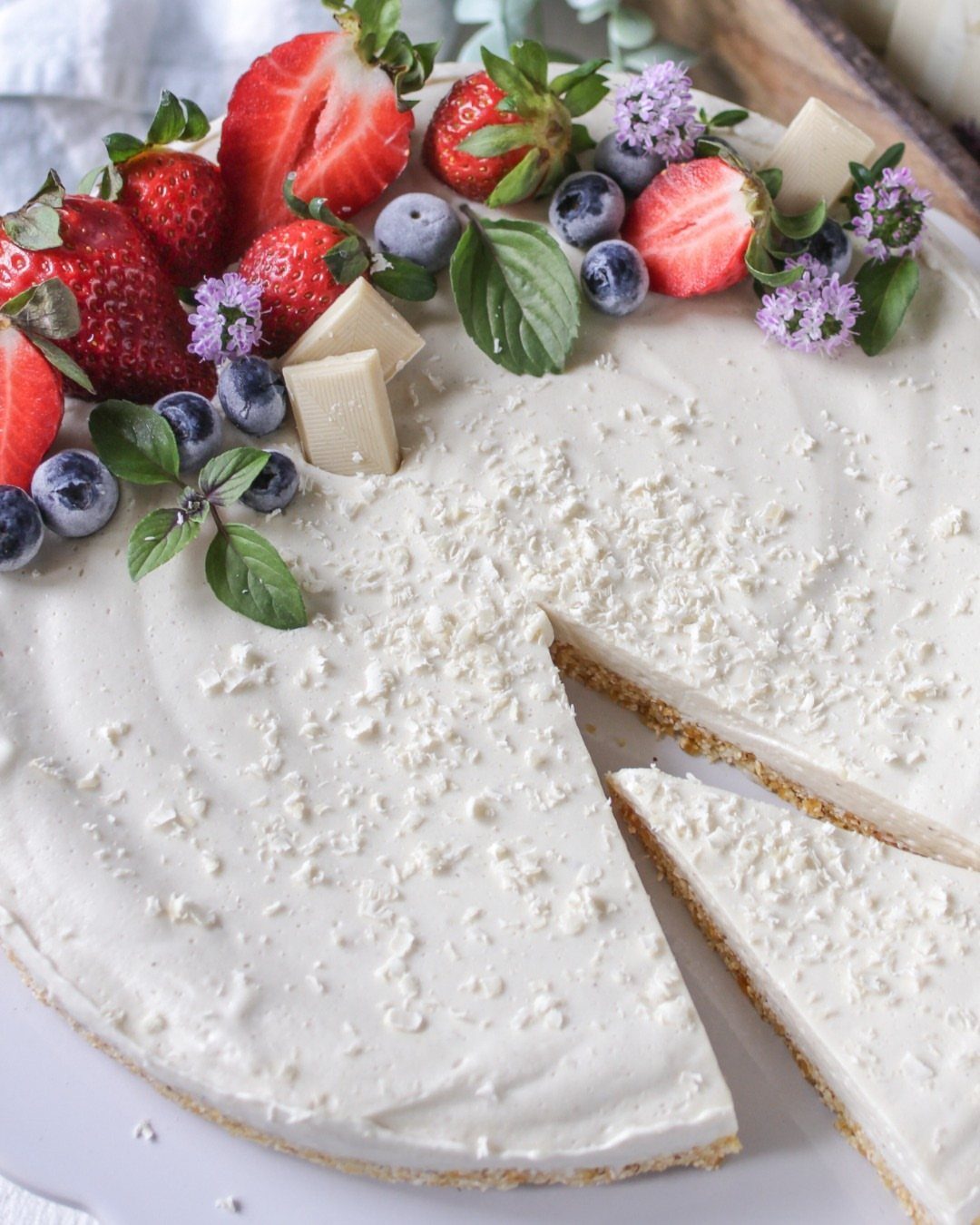 White frosted cake topped with strawberries, blueberries, white chocolate chunks, and mint leaves.