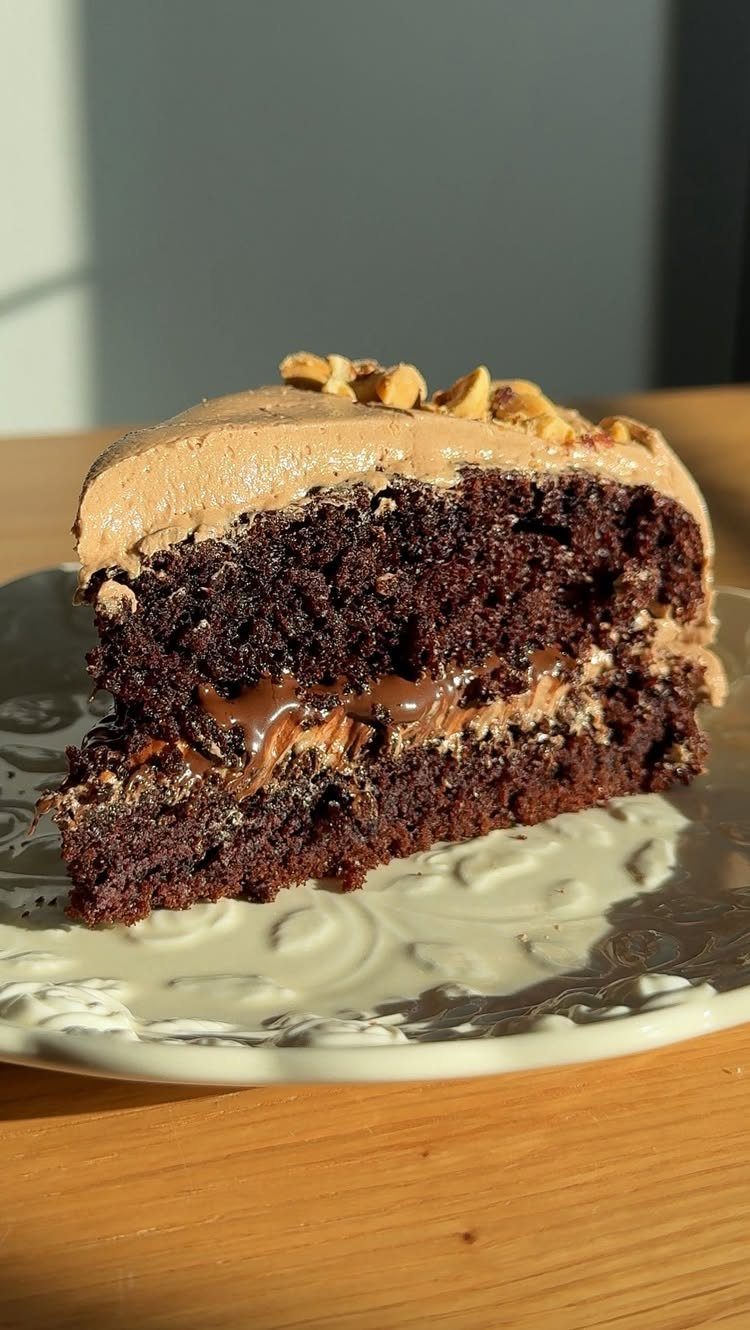 Chocolate cake slice with brown frosting, hazelnut topping, and caramel filling on a plate.