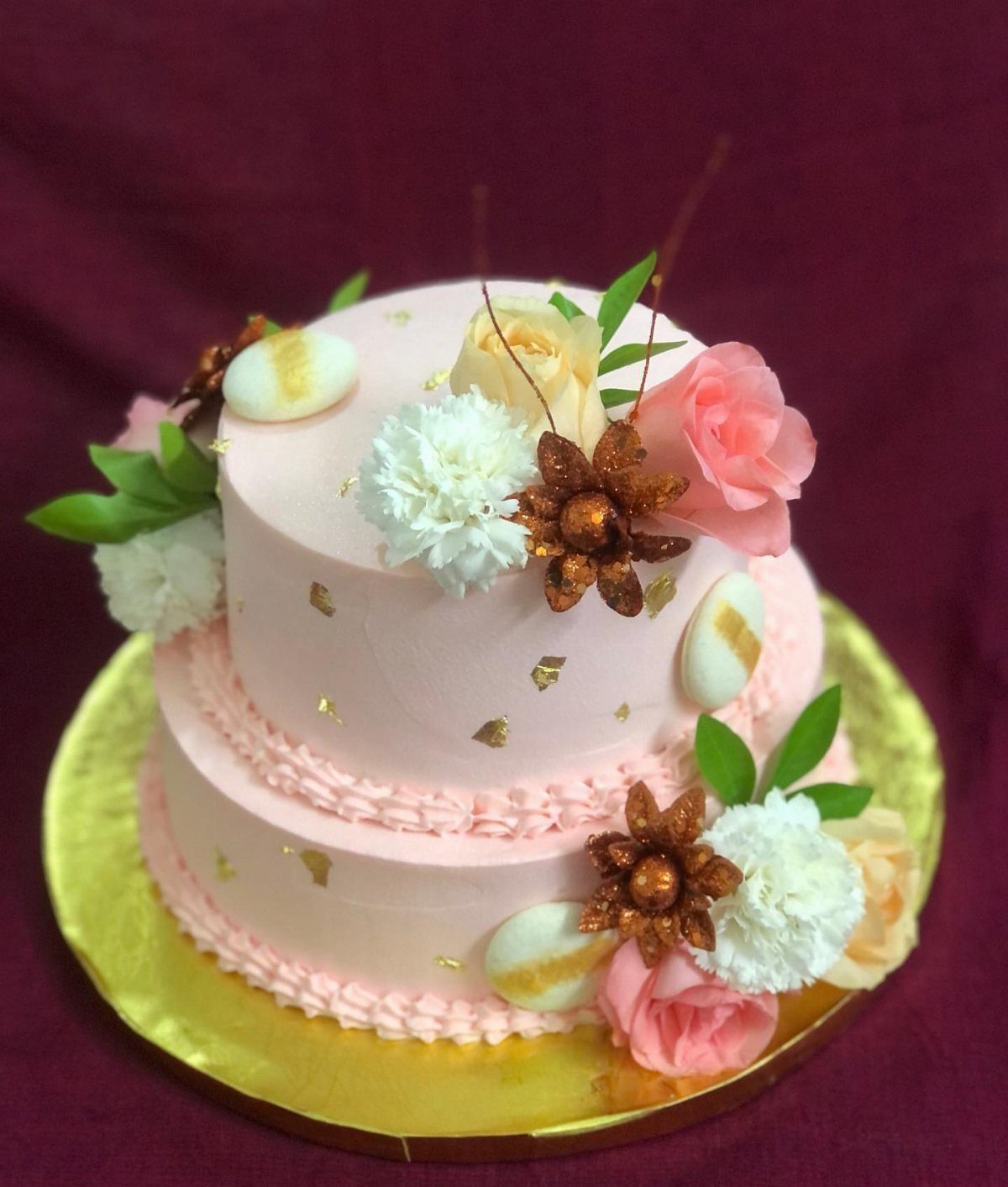 Two-tier pink frosted cake decorated with flowers and gold accents on gold board.