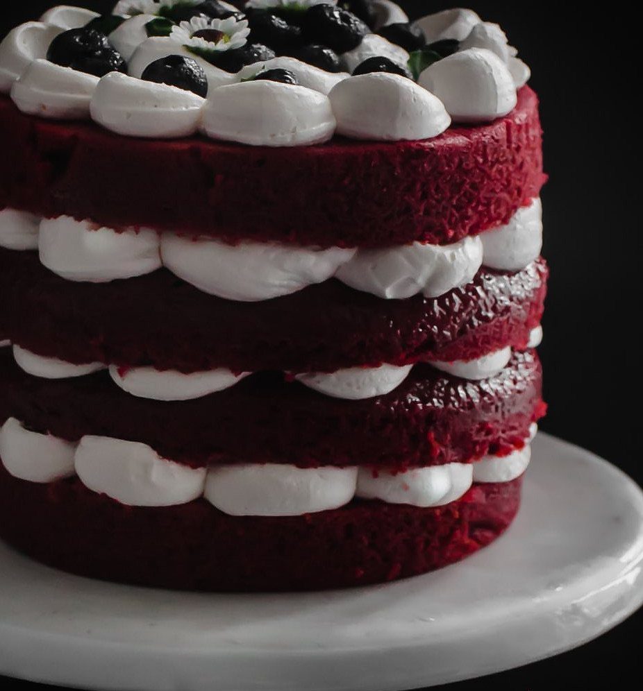 Red velvet cake with white frosting, blueberries, and flowers on a marble stand.