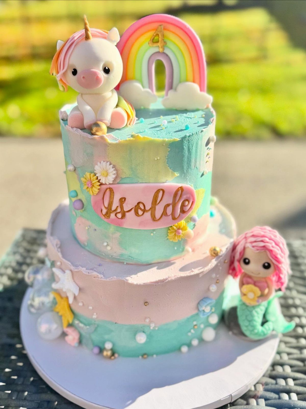 Two-tier birthday cake with unicorn and mermaid decorations in blue and pink frosting.